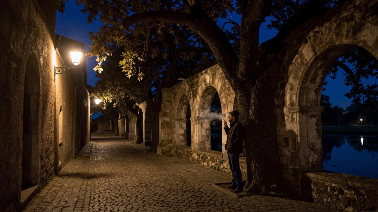 A man stands beneath a tree at midnight, smoking quietly in a cobblestone alley.